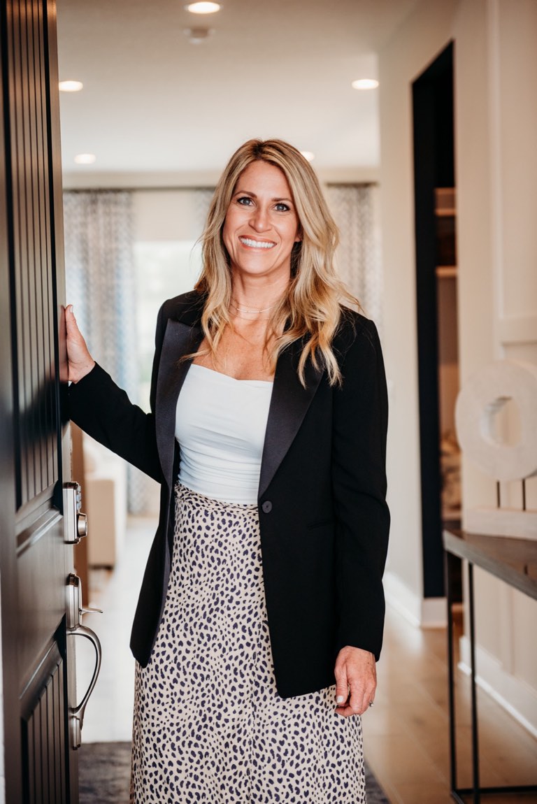 Becky Kampmeyer standing in the entry of a finished house with one hand on the edge of an open dark wood front door, wearing a black blazer over a white top and a printed skirt, with a lit hallway, patterned drapery and a console table holding a pale stone sculpture behind her