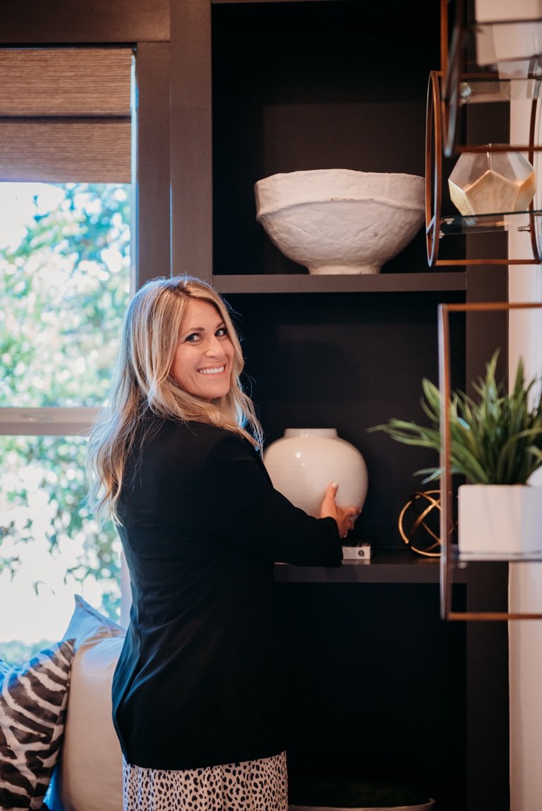 Becky Kampmeyer in a black blazer and printed skirt setting a large round white vase onto a dark built-in shelf and looking back over her shoulder, with a window under a woven shade to her left and a brass and glass etagere holding a white bowl and a potted plant to her right