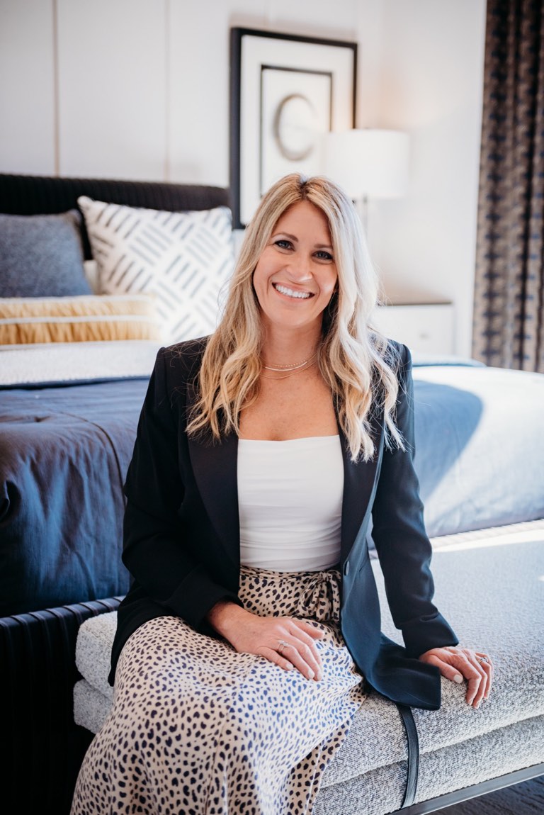 Becky Kampmeyer seated on a woven bench at the foot of a made bed in a navy and white bedroom, wearing a black blazer over a white top and a printed skirt, with a framed monogram print, a white shaded lamp and patterned curtains behind her