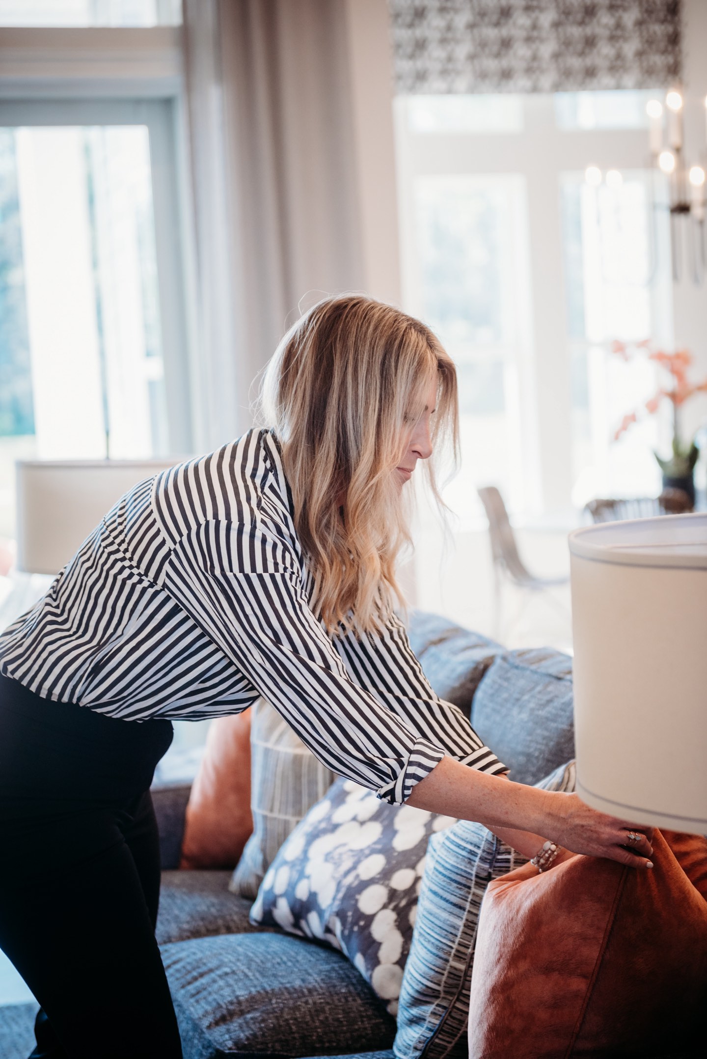 Becky Kampmeyer in a black and white striped shirt leaning over a blue-grey sofa to set a rust velvet pillow in place, with blue and white patterned cushions, a cream drum lampshade and tall sheer-curtained windows behind her