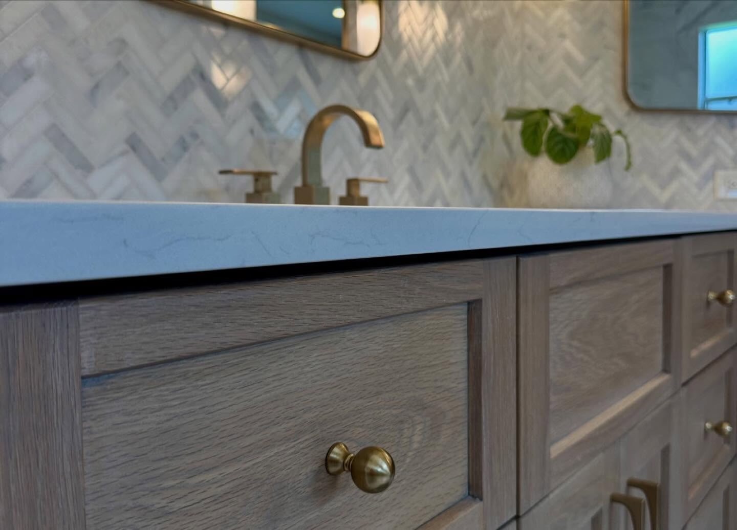 Bathroom vanity with gray-washed rift oak drawer fronts and round brass knobs under a white quartz countertop with faint gray veining, a brass widespread faucet with square lever handles, white and gray marble herringbone tile backsplash, two brass-framed mirrors, and a trailing green plant in a stone pot