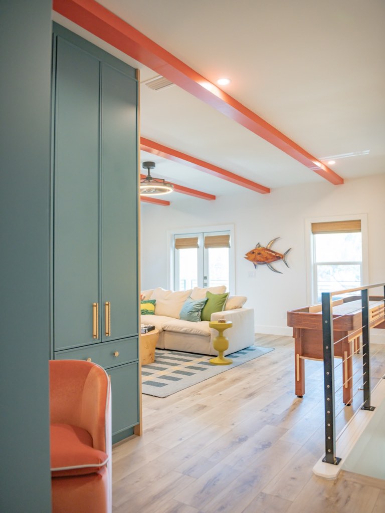 Upstairs loft with painted coral ceiling beams over a white ceiling, a floor-to-ceiling teal-green cabinet wall with slim brass pulls, a coral velvet armchair, a cream sectional on a black and white striped rug, a wood shuffleboard table and a black post and cable stair rail
