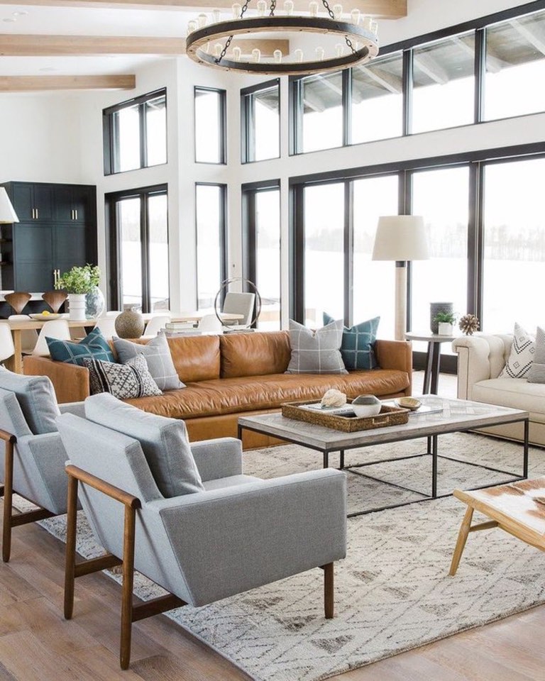 Double-height living room with a wall of black-framed windows and clerestory glass above, white oak ceiling beams and a large round wood-and-iron chandelier with exposed bulbs, a cognac leather sofa facing two pale blue-gray armchairs across a low concrete-top coffee table on a black steel base, over a cream and gray diamond-pattern rug on wide oak flooring