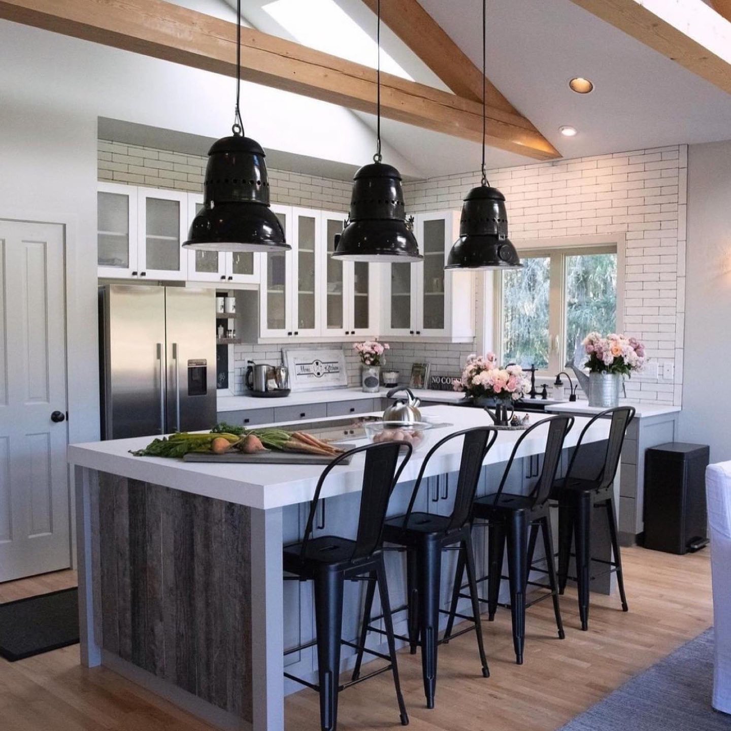 Open kitchen under a vaulted ceiling with exposed pine beams and a skylight, three oversized black enamel industrial pendants over an L-shaped island clad in weathered gray plank wood with a thick white quartz counter and four black metal stools