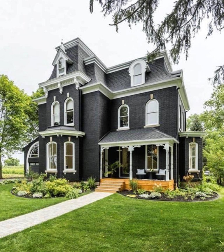 Two-story Second Empire style brick house painted black with a mansard roof, white bracketed cornice and white arched window surrounds, a covered front porch with turned white columns and scrollwork brackets sheltering two black chairs with striped cushions, new cedar steps, and a paver walkway crossing a lawn to curved beds of hostas