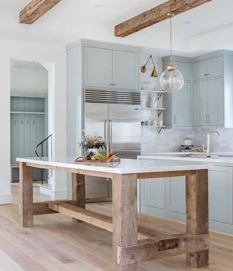Kitchen with pale sea-glass blue shaker cabinets, a reclaimed rough-sawn timber island base topped with white quartz, two rough-hewn ceiling beams, a clear glass globe pendant and stacked white open shelves against marble tile