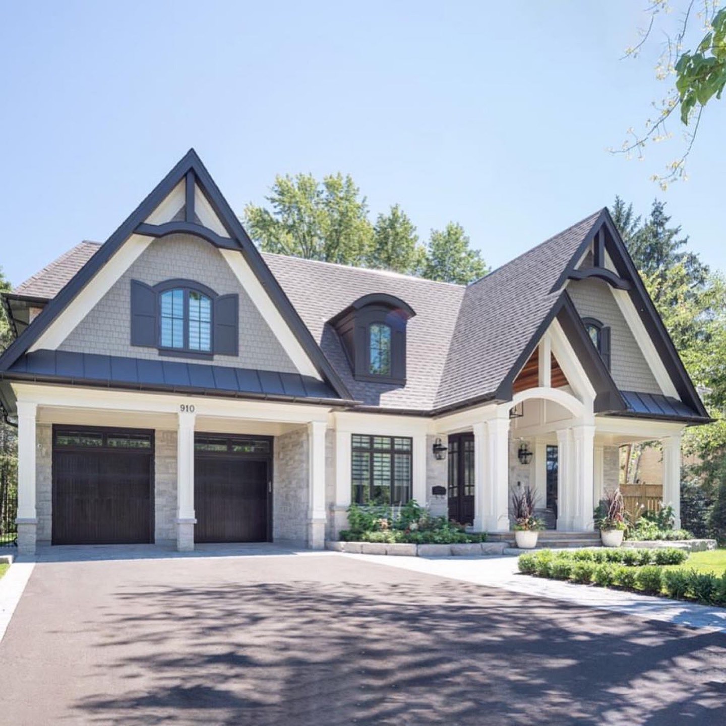 Front elevation of a two-story shingle-and-stone house with greige shingle siding, cream columns and trim, dark bronze bracketed gables, a two-bay garage with dark stained wood doors under a standing-seam metal canopy, a covered entry porch with an arched opening and lantern sconces, and clipped boxwood along a wide drive