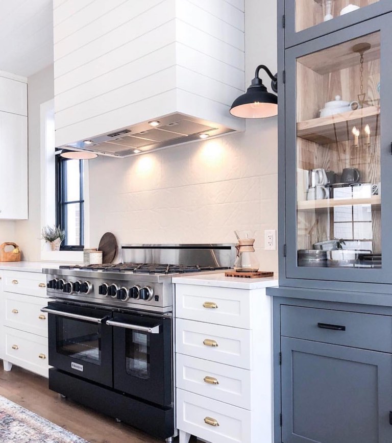Kitchen with a white shiplap-clad range hood surround over a stainless vent insert and a six-burner range, white shaker drawers with brass cup pulls, and a slate blue floor-to-ceiling glass-front cabinet with natural wood interior shelves