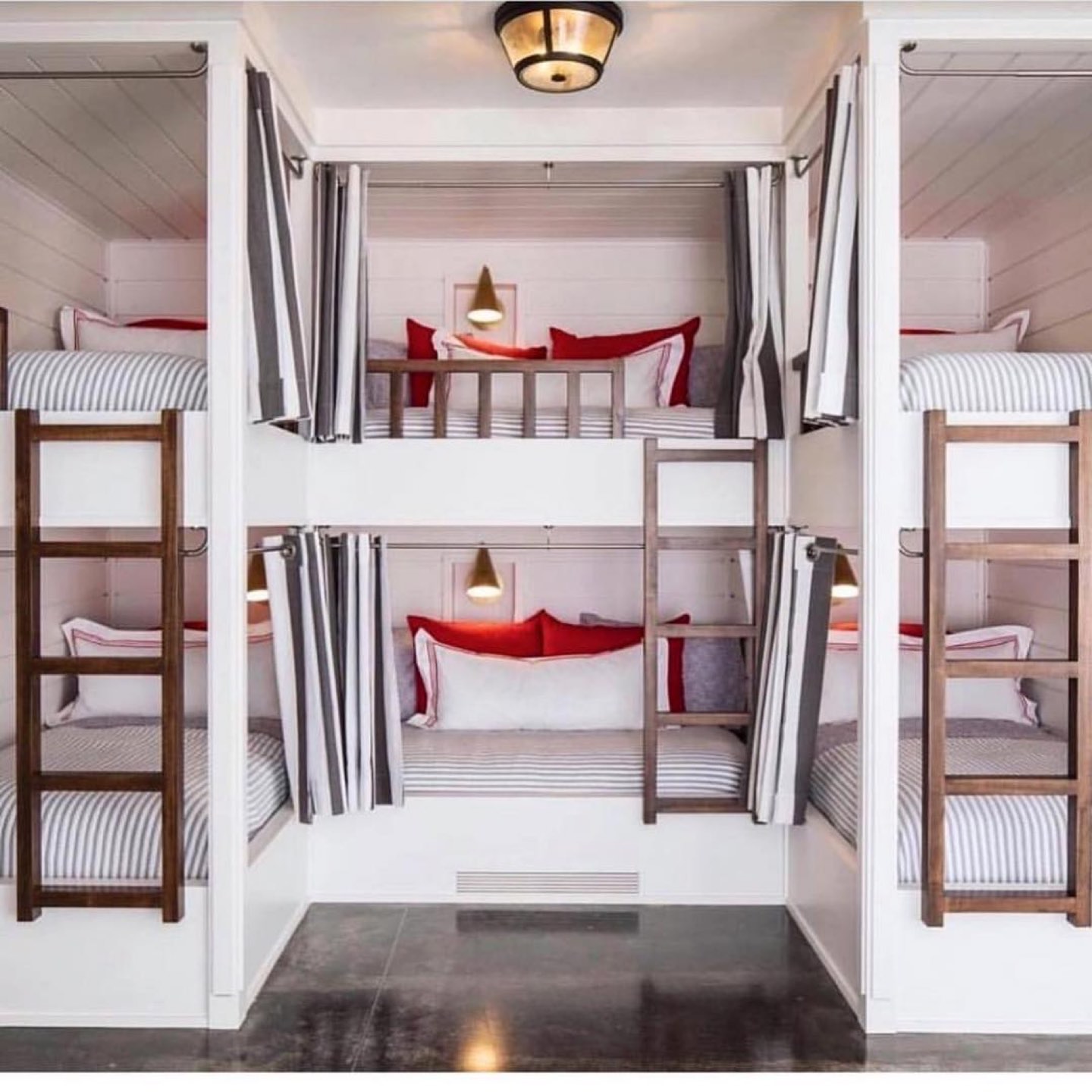 Bunk room with four built-in bunks in white shiplap alcoves, each with a ticking-stripe mattress, a brass cone sconce, striped privacy curtains and a walnut ladder to the upper berth