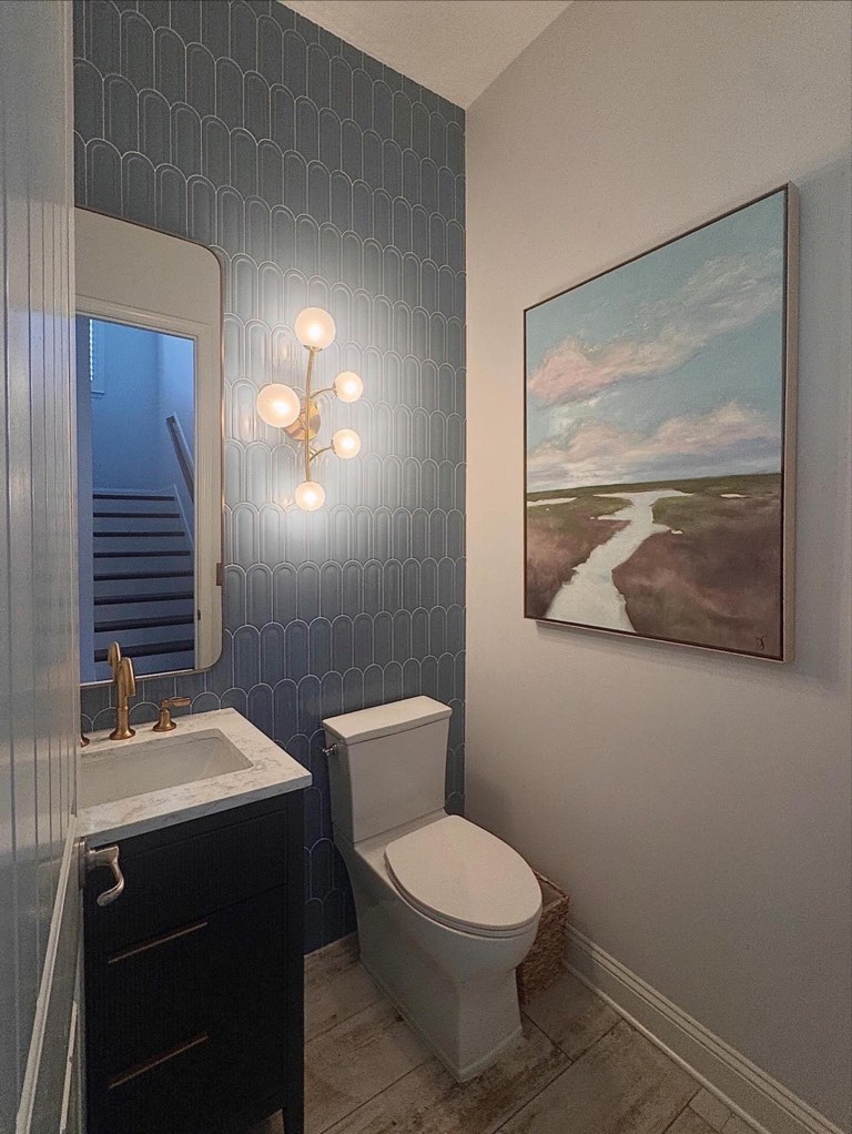 Powder room with a slate blue fan-pattern tile accent wall, a brass six-globe sputnik sconce, a navy vanity with marble countertop and brass faucet, an arched white-framed mirror and a framed marsh landscape painting on the adjacent gray wall