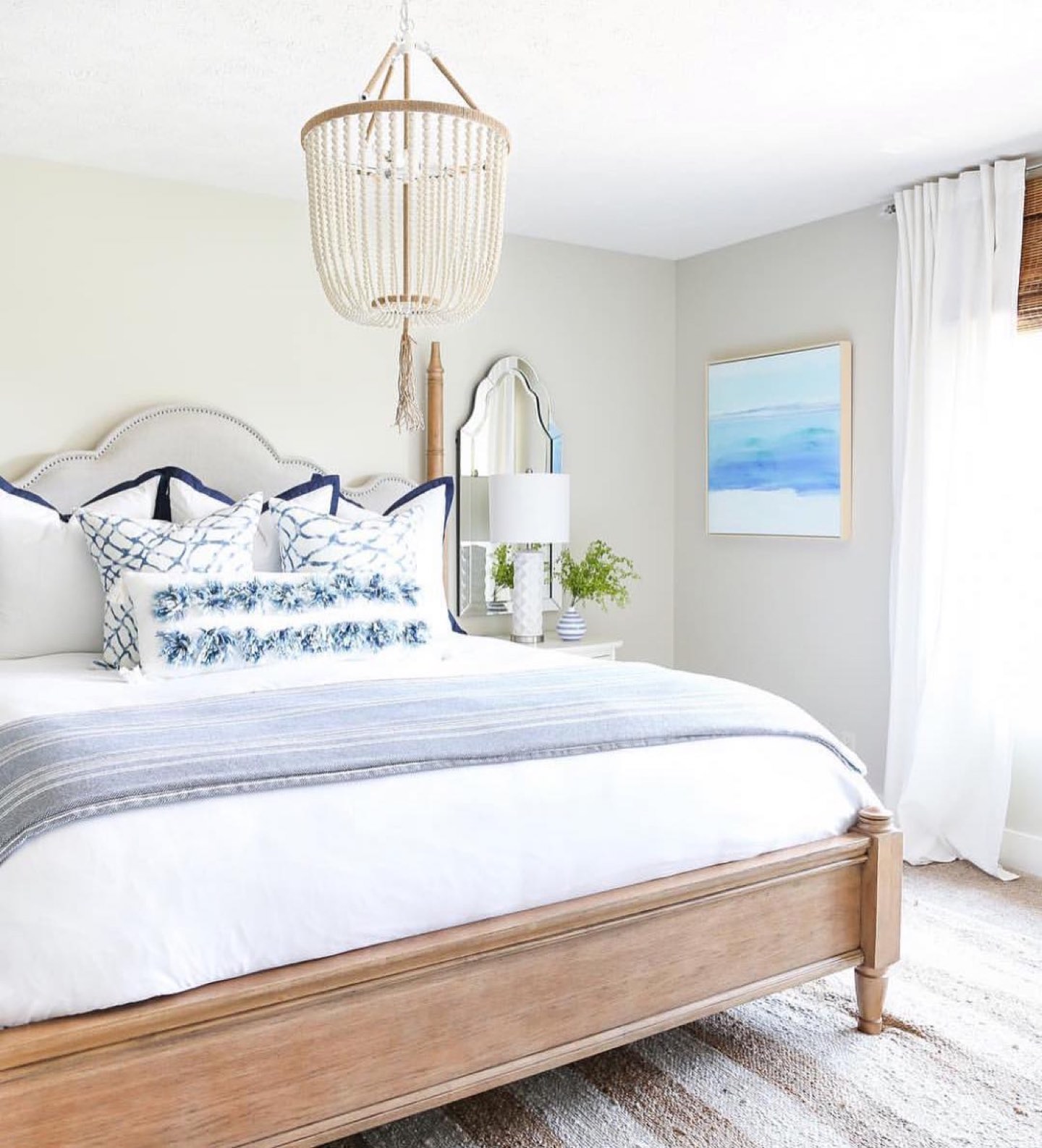Bedroom with a cream upholstered arched headboard with nailhead trim on a light oak bed frame, white bedding layered with a blue and white striped throw and indigo net-print pillows, a white beaded chandelier above the bed, an arched mirror and white ceramic lamp on the nightstand, and a jute rug on the floor