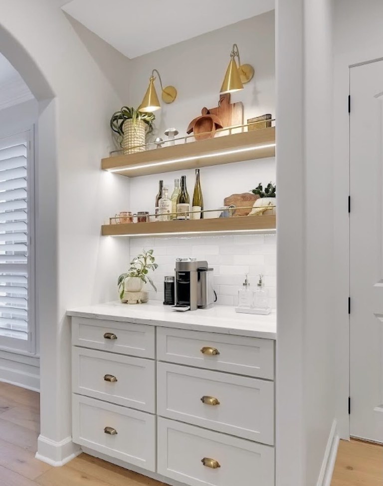 Built-in coffee bar niche with white shaker drawers and brass cup pulls, white quartz counter, stacked white subway tile backsplash, two white oak floating shelves with brass gallery rails and integrated LED strips, and two brass cone wall sconces