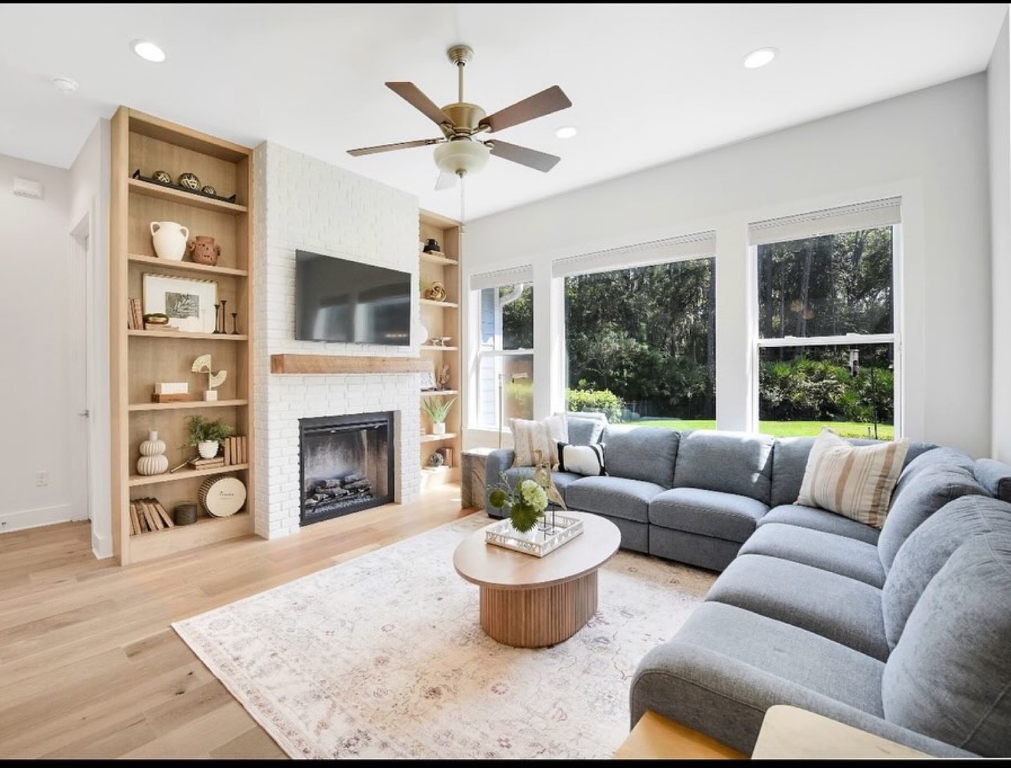 Living room with a painted white brick fireplace and raw oak mantel flanked by light oak open built-in shelving, a blue-gray channel-tufted sectional, round fluted oak coffee table and wide-plank oak floors
