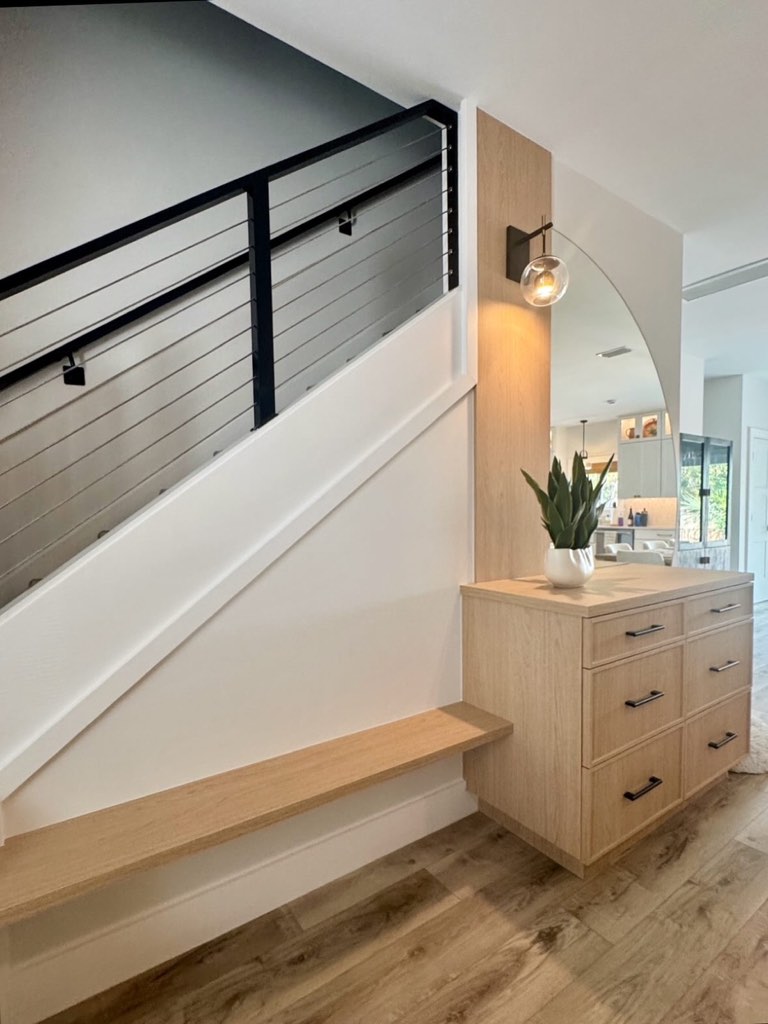 Stair landing with a floating white oak bench built into a white shiplap stair wall, a black metal and horizontal cable railing above, a six-drawer white oak cabinet with matte black bar pulls, an arched frameless mirror on a white oak panel, and a black-arm sconce with a clear glass globe