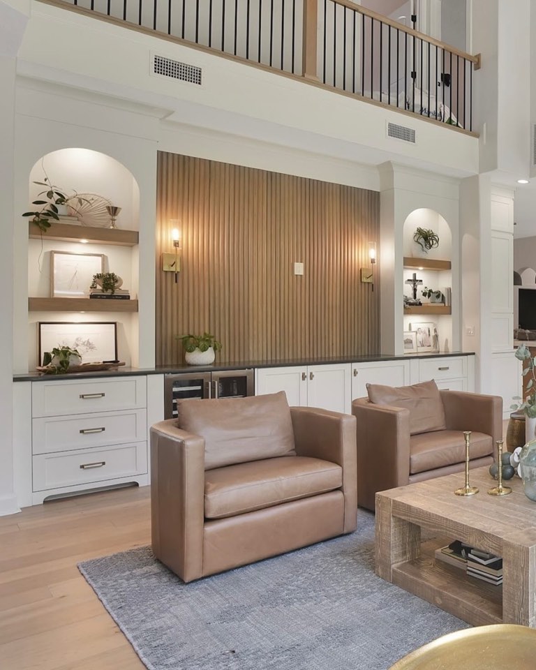 Two-story living room with a fluted oak slat feature wall between two lit arched niches with floating oak shelves, white shaker cabinetry topped with a dark stone counter and a built-in beverage fridge, brass plate sconces, two camel leather swivel chairs on a pale blue-gray rug, a chunky reclaimed wood coffee table with brass candlesticks, light oak flooring, and a black spindle railing on the balcony above