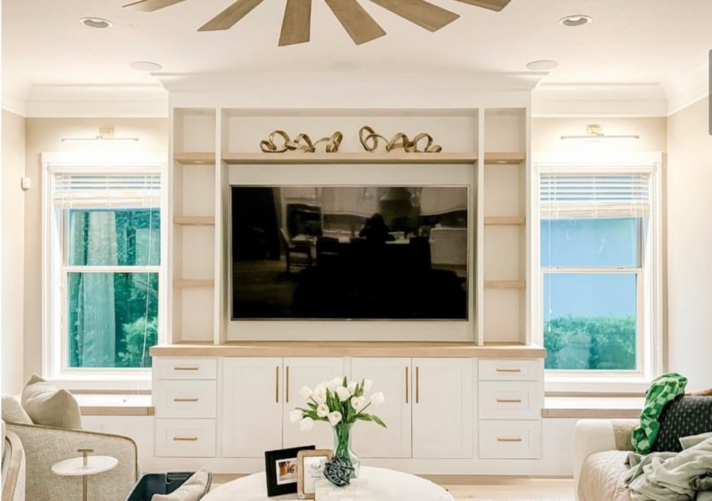 Living room built-in media wall in painted white with light oak floating shelves and counter, a wall-mounted television in a recessed surround, a brass ribbon sculpture on the upper shelf, flat-panel cabinet doors with slim brass pulls, flanking double-hung windows with white blinds and brass picture lights, a wood-blade ceiling fan, a glass vase of white tulips on a round table, and an upholstered cream armchair at left