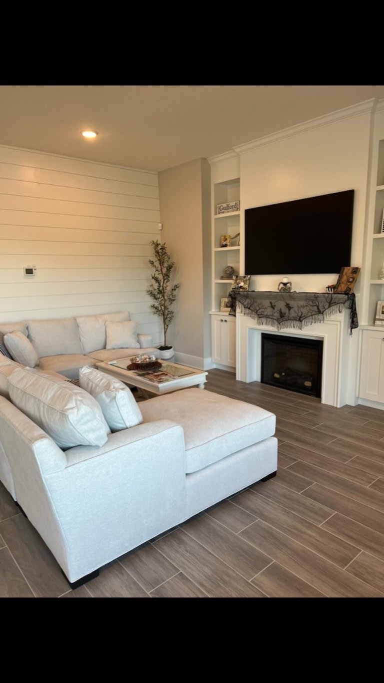 Living room with a cream chenille sectional and chaise on gray wood-look plank tile, a horizontal shiplap accent wall, white built-in cabinets and open shelves flanking a white mantel fireplace with a wall-mounted television above, and a potted olive tree in the corner