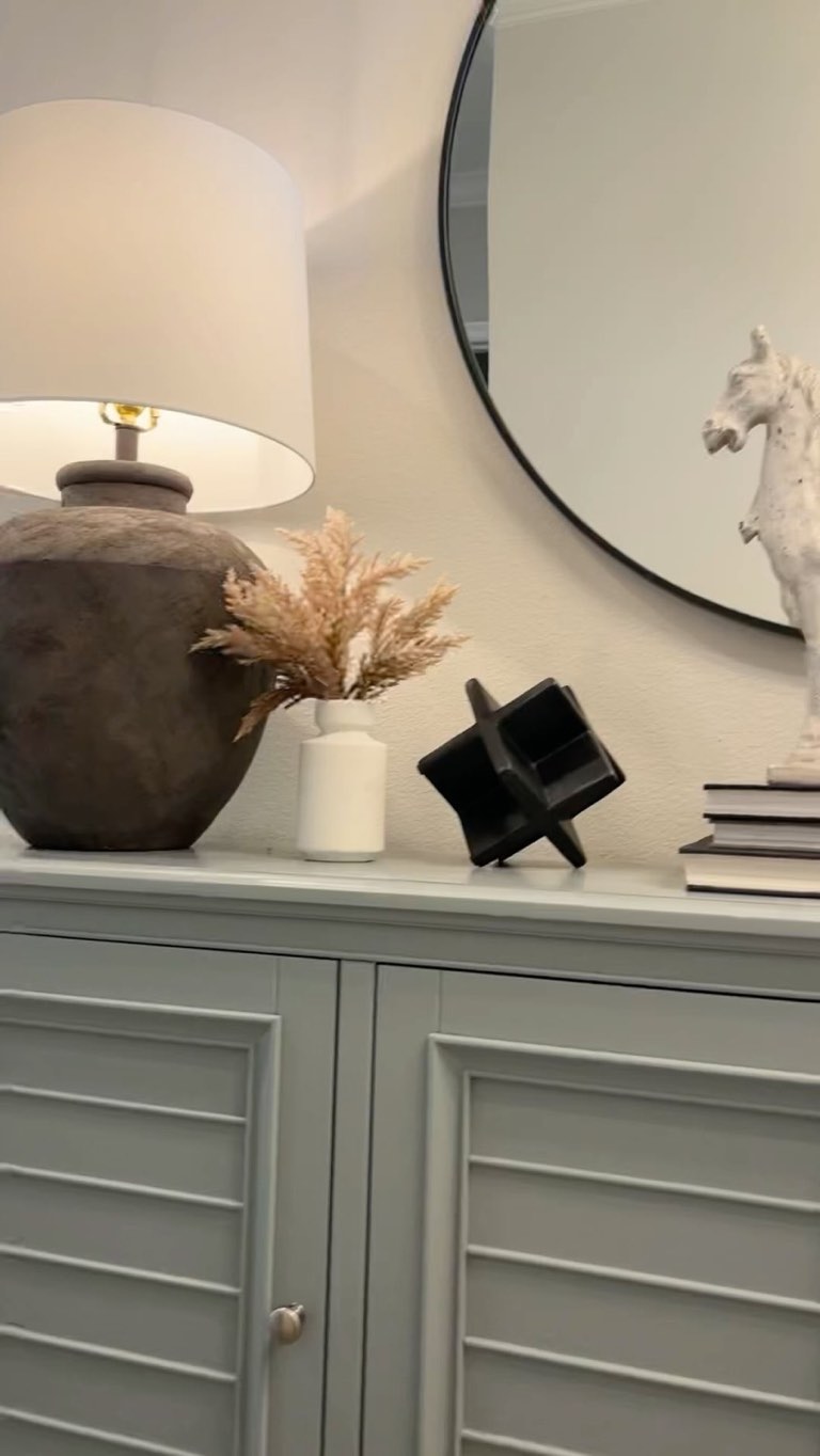Styled vignette on a sage green louvered cabinet with a matte terracotta urn lamp and white drum shade, dried pampas in a small white vase, a black sculptural cross-form object and a stack of white books beneath a round black-framed mirror