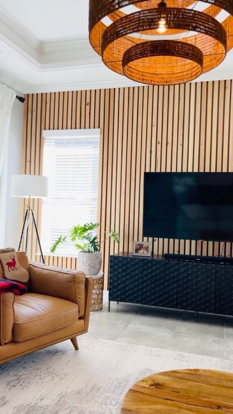 Living room with a floor-to-ceiling vertical wood slat accent wall behind a wall-mounted TV, a black textured three-door media console, tan leather mid-century armchair, woven rattan double-tier drum chandelier, white tripod floor lamp, split-leaf philodendron in a white pot, faded gray area rug over gray tile, and a live-edge wood coffee table