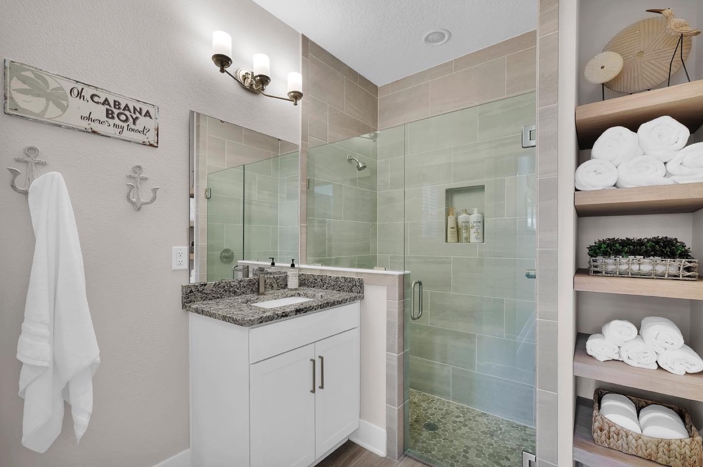 Guest bathroom with a white shaker vanity under a speckled granite counter, a walk-in glass shower tiled in sage green with a green pebble floor and a recessed niche, and open wood shelves stacked with rolled white towels