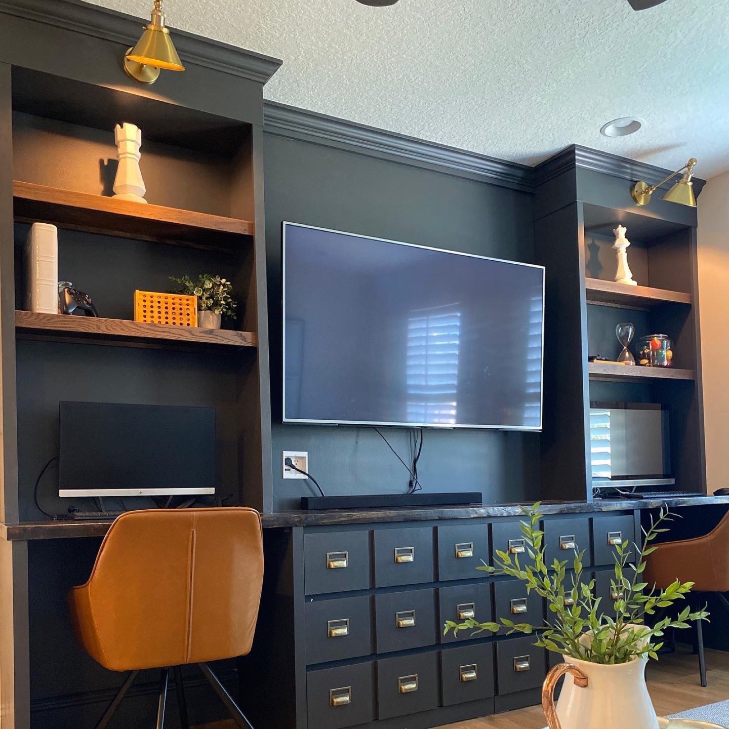 Built-in home office and media wall painted dark charcoal green with stained oak shelves, brass swing-arm and cone sconces, a wall-mounted TV over an apothecary-style bank of drawers with brass label pulls, a live-edge dark wood desk ledge, two cognac leather swivel chairs, and a white pitcher of greenery