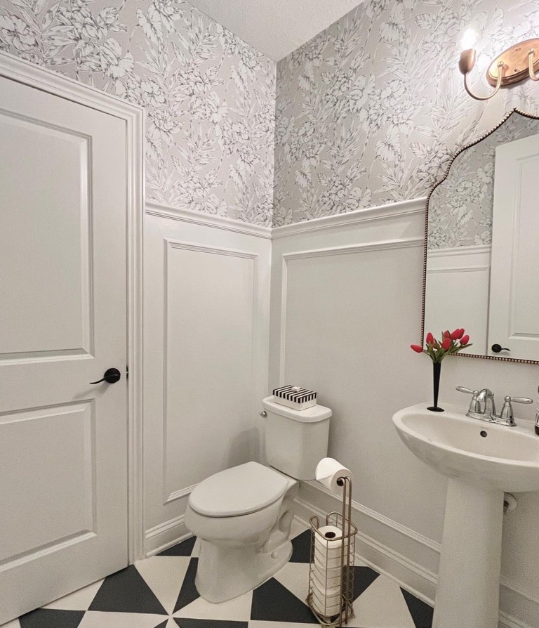 Powder room with gray and white floral toile wallpaper above white picture-frame wainscoting, a black and white harlequin tile floor, a white pedestal sink with a chrome widespread faucet, an arched copper beaded mirror, a copper two-arm sconce, and black door hardware