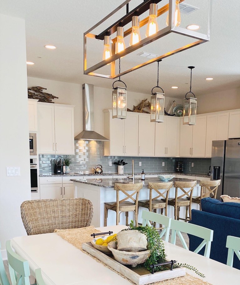 Open coastal kitchen with cream shaker cabinets, blue-gray glass subway backsplash, speckled granite counters, three lantern pendants over the island and four X-back wood counter stools