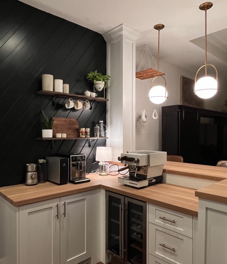 Corner coffee bar with a diagonal black-painted shiplap wall, two walnut floating shelves on black iron brackets holding ceramic canisters and hanging mugs, butcher-block counters over cream shaker cabinets, an espresso machine and a glass-front wine fridge