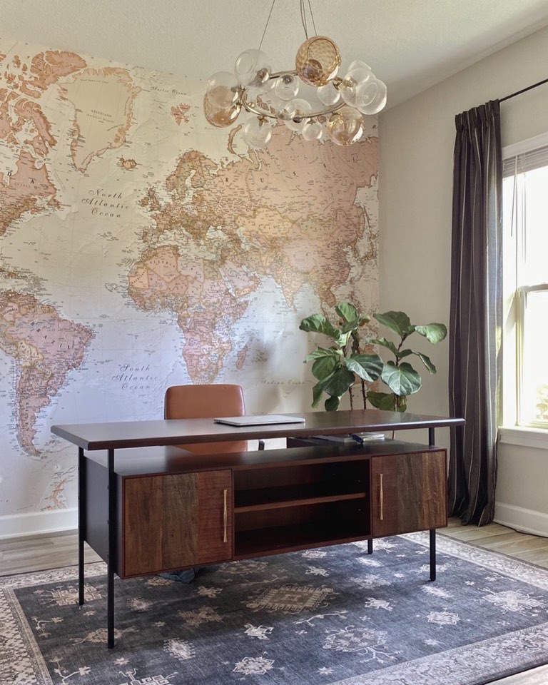 Home office with a full-wall vintage world map mural in sepia and blush tones, a walnut and black steel desk with a credenza of live-edge wood doors and slim brass pulls, a cognac leather task chair, a brass chandelier with clustered clear and amber glass globes, and a faded slate-blue Persian rug