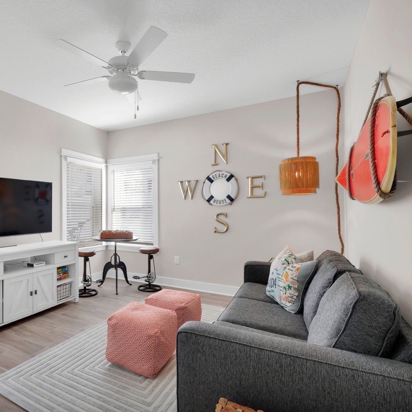 Beach-house bonus room with a charcoal fabric sofa, two coral geometric-print floor poufs on a striped ivory rug, a white barn-door media console under a wall-mounted television, a black cafe table with two industrial crank stools in the window bay, gold compass-point letters around a nautical life ring on the wall, a fringed rattan plug-in pendant on a rope cord, and a coral surfboard mounted on rope hangers