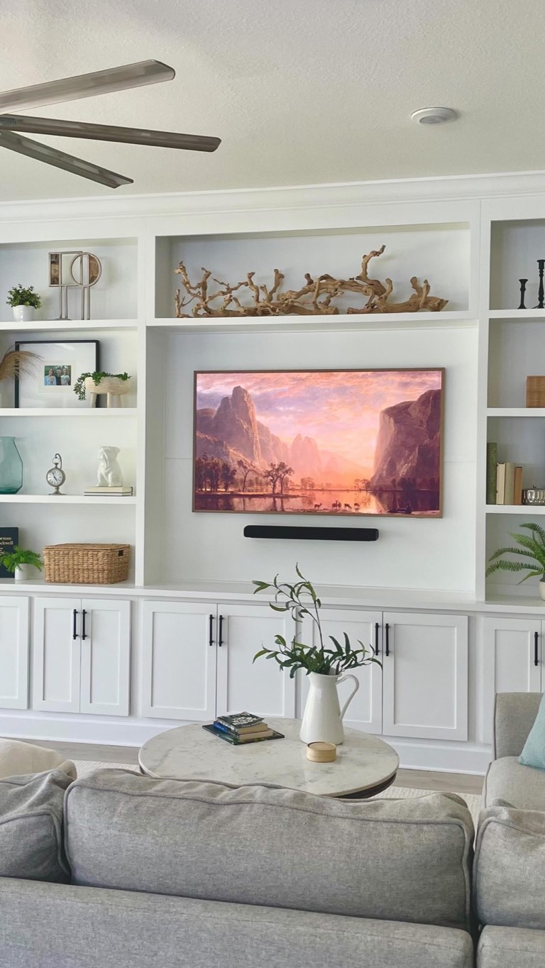 Living room wall of white built-ins with open shelves and shaker cabinet doors with matte black bar pulls, flanking a recessed television in a wood frame, with a gray upholstered sofa and a round marble coffee table in the foreground
