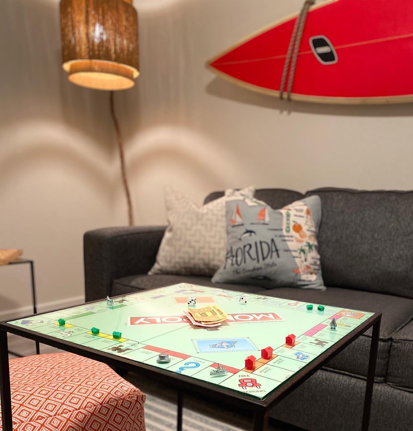 Game lounge with a charcoal sofa, a Monopoly board set up on a black metal coffee table, a red and white surfboard mounted on the cream wall, a woven wood drum pendant and a coral geometric ottoman on a striped rug