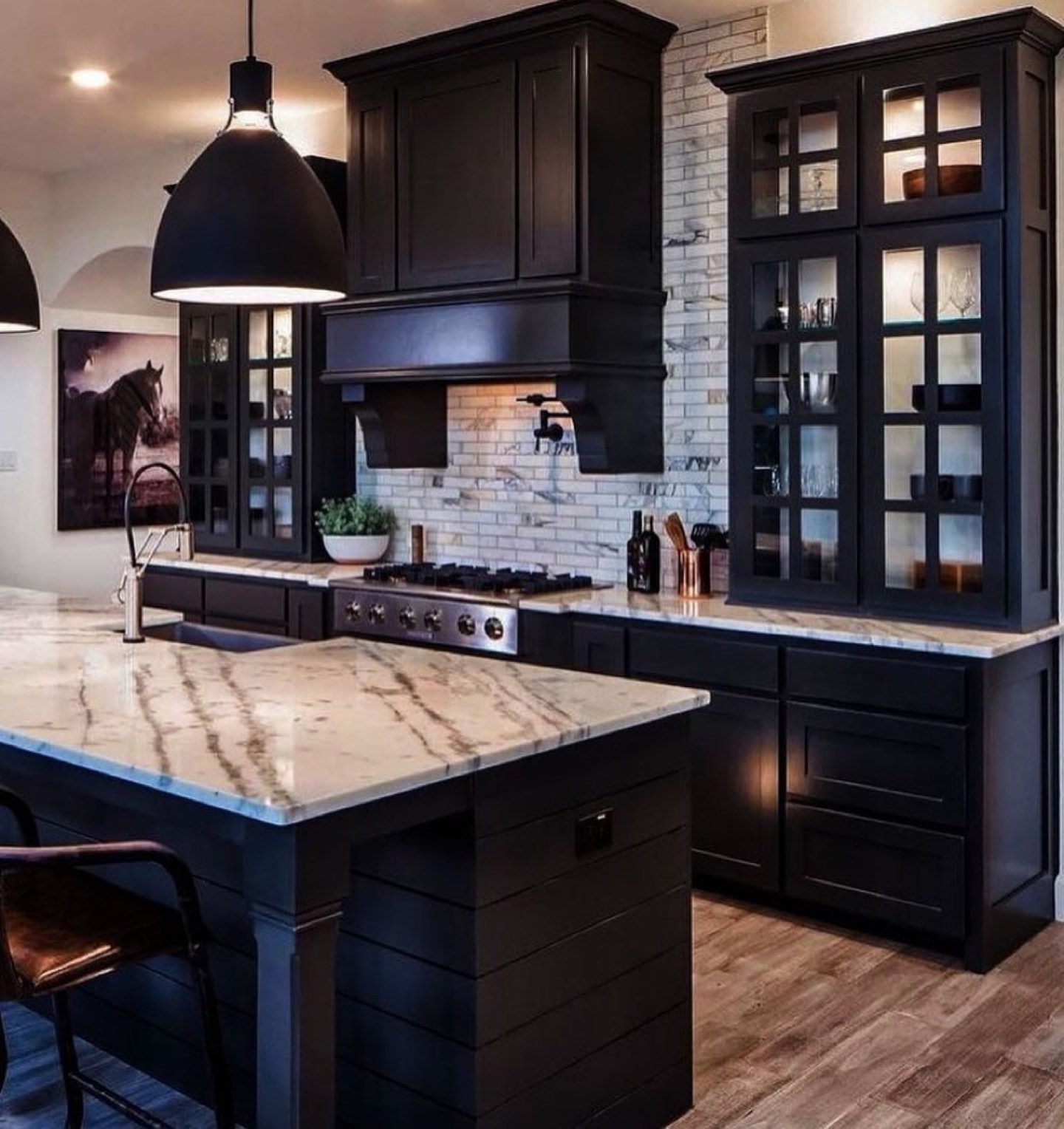 Kitchen with black shaker cabinetry, tall glass-front lit display cabinets, a white marble waterfall island and counters, a marble subway backsplash behind a stainless gas rangetop with a black hood, a black dome pendant, and wide-plank wood floors