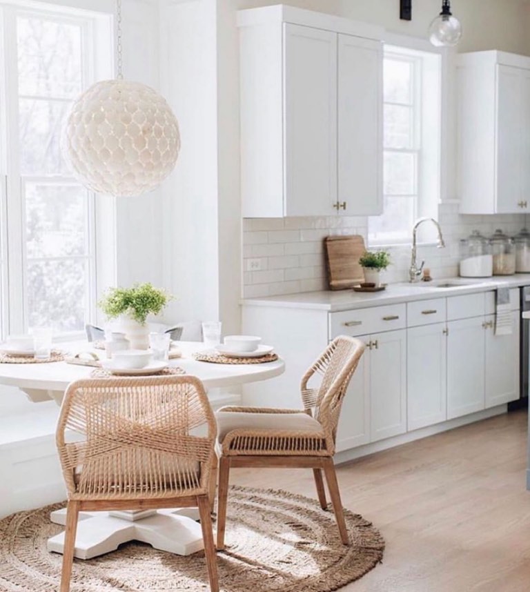 White kitchen with shaker cabinets, brass bar pulls, a white subway tile backsplash and a stainless gooseneck faucet, opening to a breakfast nook with a round white pedestal table, woven rope-back chairs, a round jute rug and a white capiz globe pendant