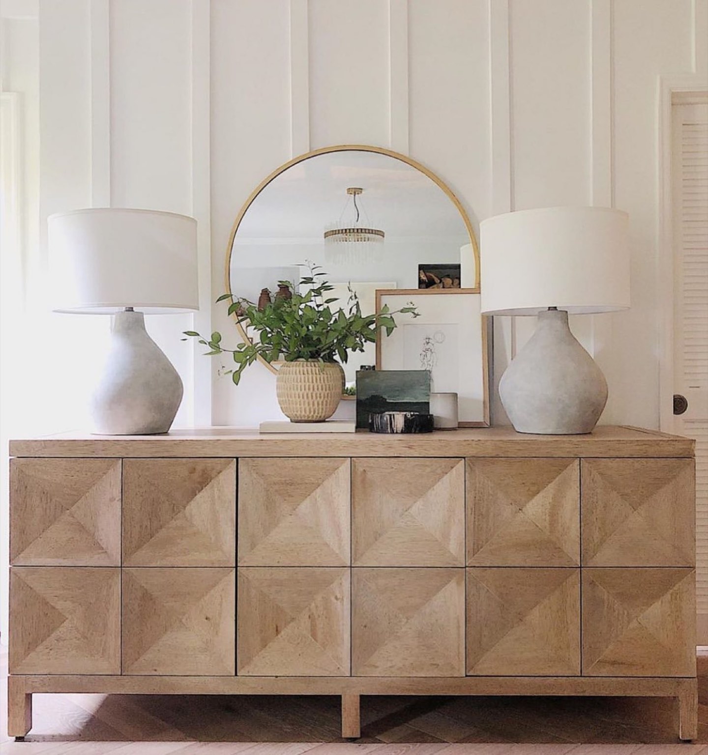 Bleached oak sideboard with faceted diamond-front doors against a board-and-batten white wall, flanked by two concrete gourd lamps with white drum shades, with an arched brass-framed mirror, a ribbed ceramic vase of green foliage and small leaning artwork
