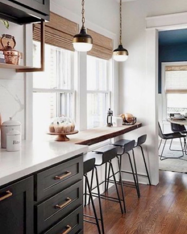 Kitchen with dark charcoal-green shaker drawer fronts and aged brass bar pulls under a white quartz counter, a live-edge wood plank set into the windowsill as a bar with three black metal and leather stools, two black and brass globe pendants, and woven bamboo Roman shades
