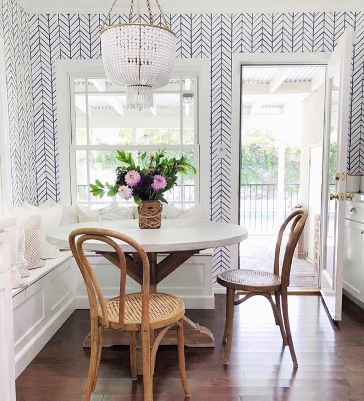 Breakfast nook papered in a navy hand-drawn herringbone pattern on white, with a white beaded chandelier on a brass frame over a round white-topped trestle table, a built-in white banquette lining two walls, two bentwood chairs with cane seats, and an open French door onto a covered patio and pool