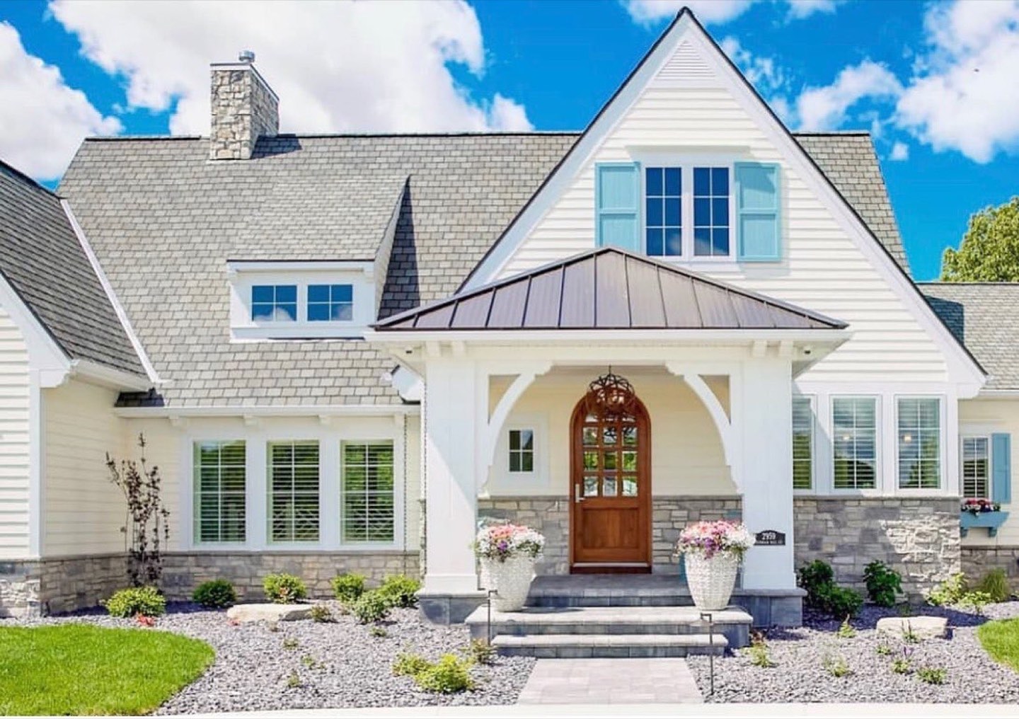Front elevation of a white lap-sided house with a gray shingled roof, stone foundation veneer and a stone chimney, a gabled entry porch with a bronze standing-seam roof over an arched stained-wood front door, pale blue shutters on the upper dormers and white urn planters flanking the bluestone steps
