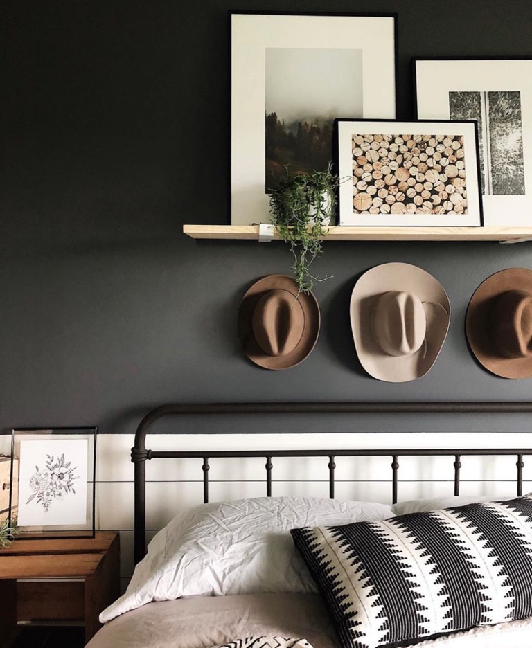 Bedroom with a charcoal painted wall above white shiplap wainscot, a light wood picture ledge holding framed prints and a trailing potted plant, three tan and camel felt hats hung in a row, and a black metal spindle bed made up with white linen bedding and a black-and-white mudcloth pillow