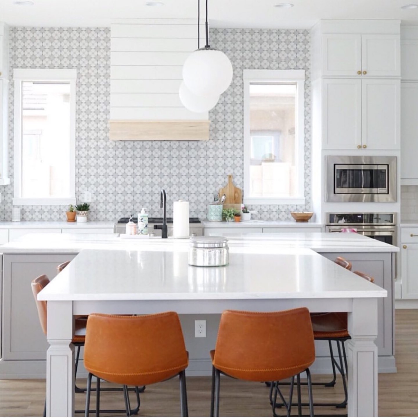 White kitchen with gray patterned encaustic-look tile covering the backsplash wall, a shiplap range hood banded in light oak, a milk glass globe pendant, white shaker cabinets with brass knobs, and a white quartz island that extends into a table with turned legs and four cognac leather barrel stools