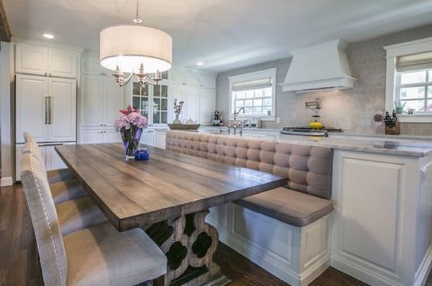 Kitchen with white raised-panel cabinetry and a plaster-look vent hood, gray marble counters, and a taupe tufted banquette wrapping the peninsula beside a weathered oak trestle table under a white drum shade chandelier