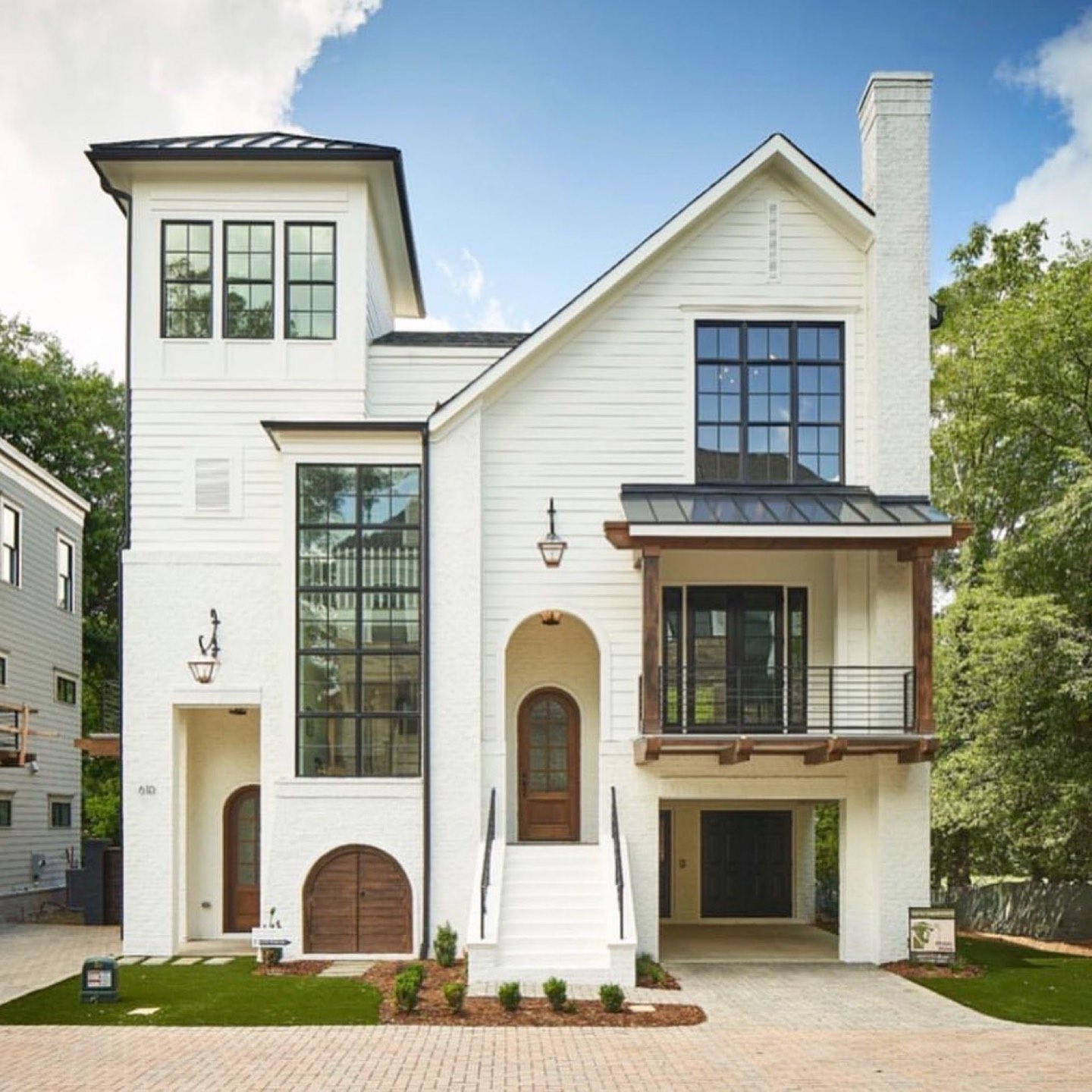 Three-story white painted brick and lap-siding house with black-framed windows, a standing-seam metal awning over a second-floor balcony with stained timber brackets, an arched walnut front door, and a brick paver drive