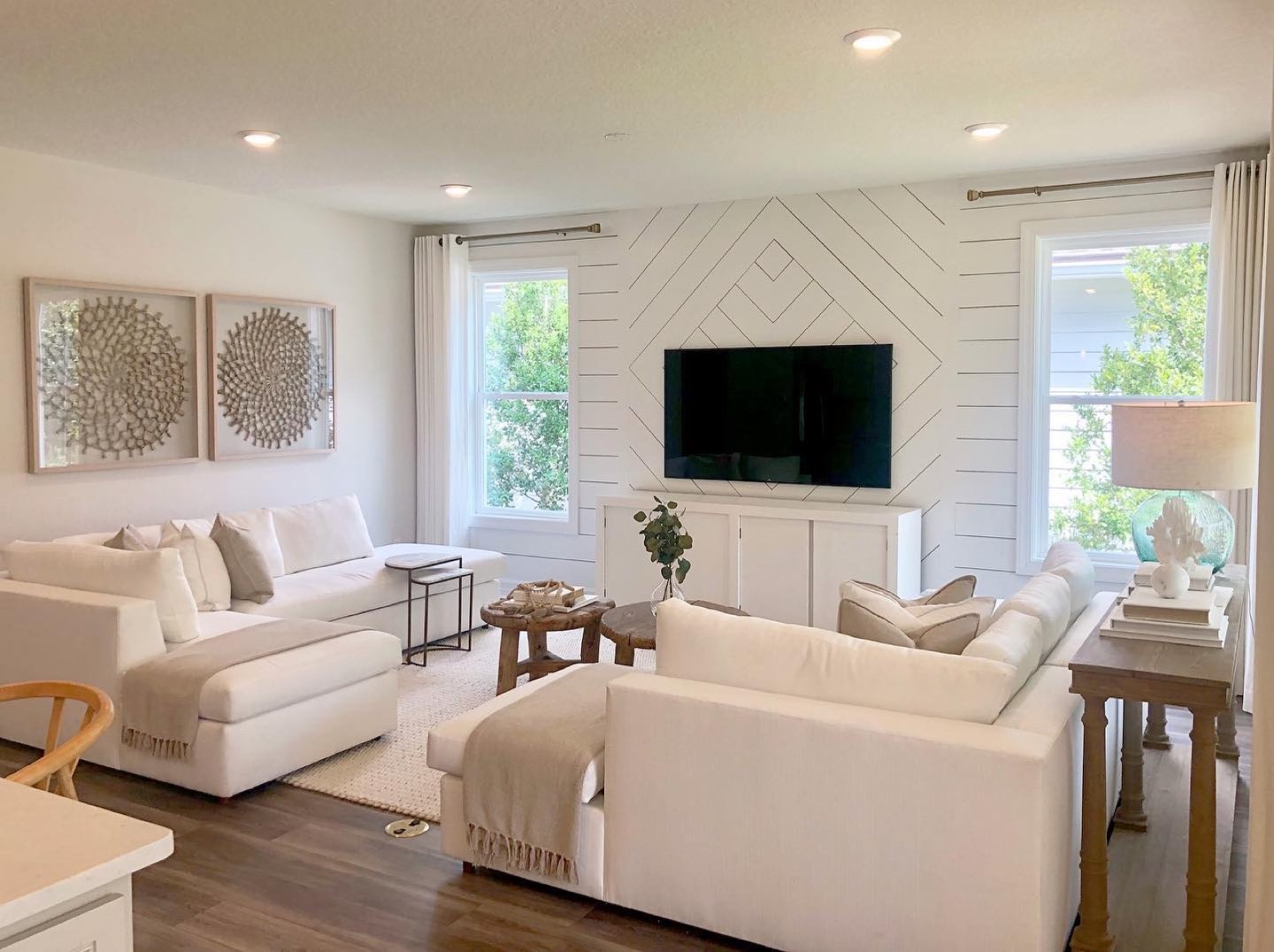 Living room with two ivory slipcovered sofas facing each other on a jute rug, a white shiplap accent wall laid in a diamond pattern behind a wall-mounted television, a low white media cabinet, round wood side tables, and dark walnut plank flooring