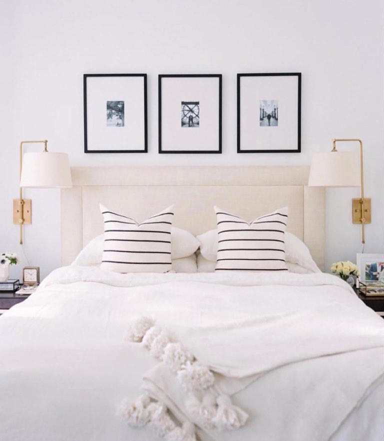 Bedroom with a cream upholstered panel headboard, brass swing-arm sconces on either side, three black-framed black-and-white photographs hung above, white linen bedding with black-striped square pillows and a pom-pom trimmed throw