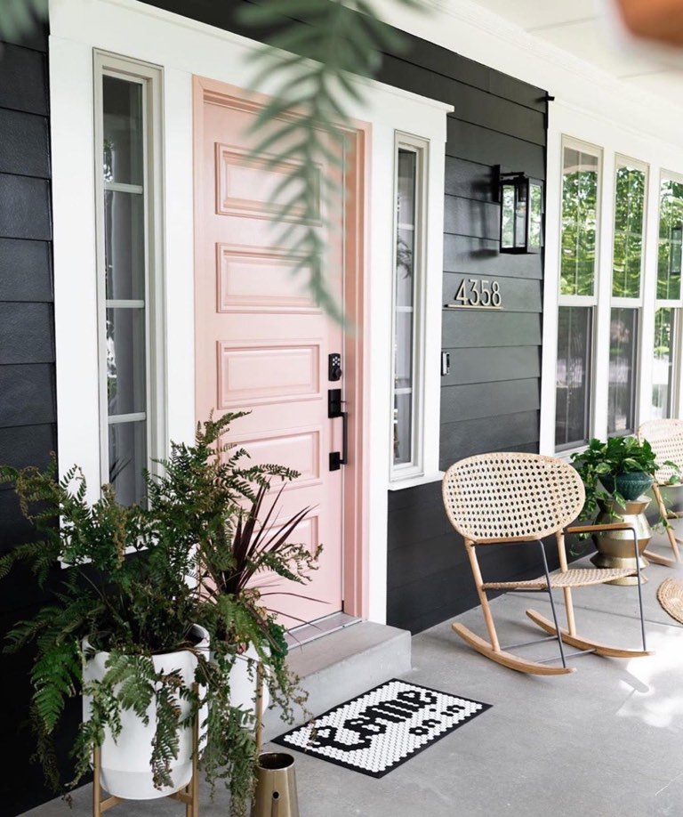 Covered front porch of a black lap-siding house with white trim, a blush pink five-panel front door flanked by narrow sidelights, a black lantern sconce, brass house numbers, cane-back rocking chairs on a concrete slab and ferns in a white pot