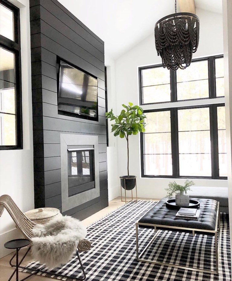 Vaulted living room with a floor-to-ceiling black shiplap fireplace wall holding a mounted television and a steel-surround firebox, black-framed windows, a dark wood bead chandelier, a black-and-white buffalo check rug, and a tufted black leather ottoman on a brass frame