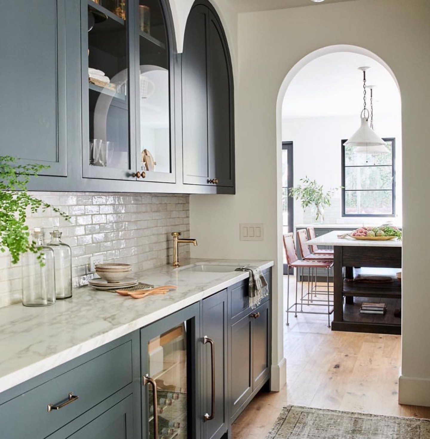 Butler's pantry with slate blue-green shaker cabinets, arched glass-front upper doors, white marble countertop and glazed white subway tile, a brass gooseneck faucet over an undermount sink, a glass-front wine fridge in the lower run, and a plaster arched doorway opening to a dining area with a dark wood island and white cone pendants