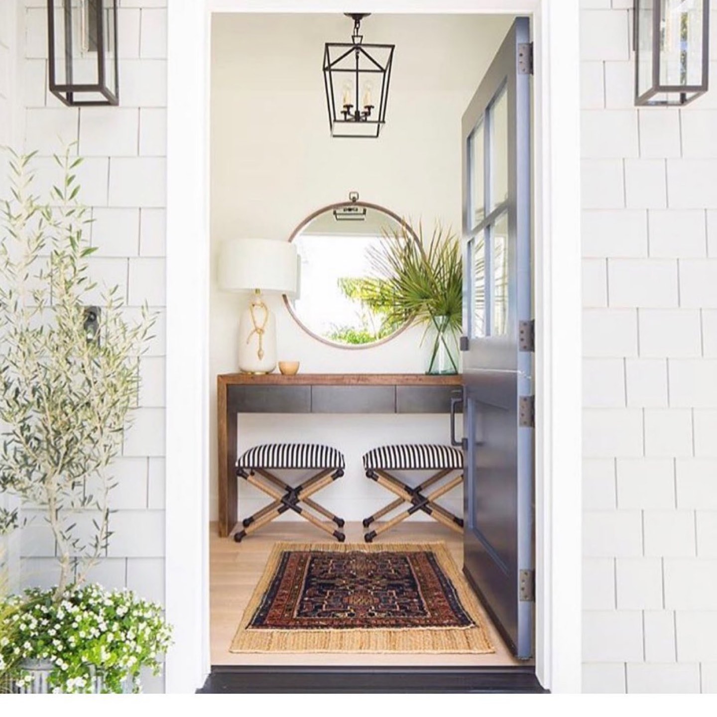 Entry seen through an open dark gray glass-paned door flanked by two black lantern sconces, with a walnut waterfall console holding a white ceramic lamp and a glass vase of palm fronds beneath a round bronze-framed mirror, two navy and white striped X-base stools with rope-wrapped legs, and a Persian rug layered over jute