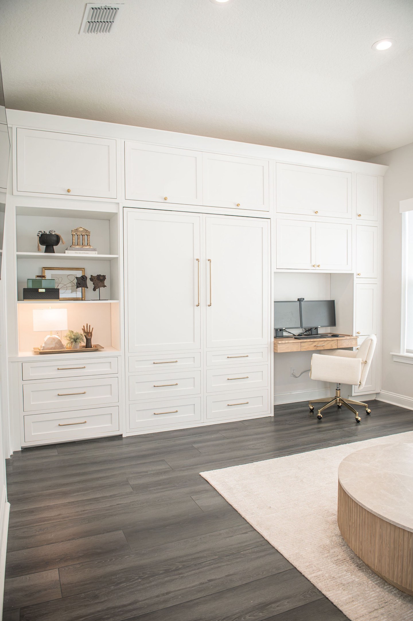 Full wall of white built-in cabinetry with brass hardware, the Murphy bed closed flush behind panelled doors, a lit open niche styled with books and objects, and a white oak desk with dual monitors and a cream upholstered chair at the far end