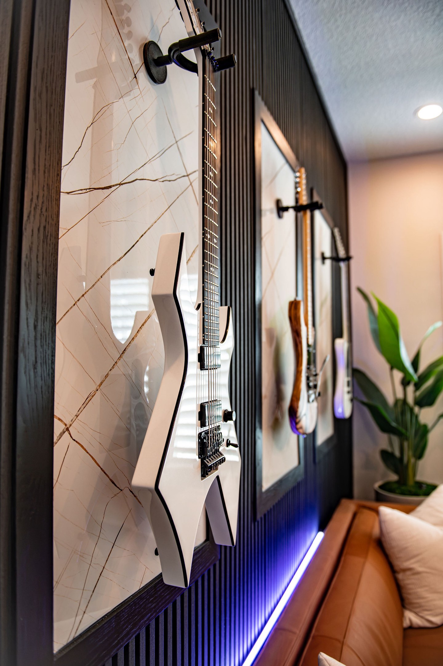 Close view along a black fluted wall of framed electric guitars mounted on black brackets over marble-look panels, with violet LED light washing the fluting and a cognac leather sofa and palm below